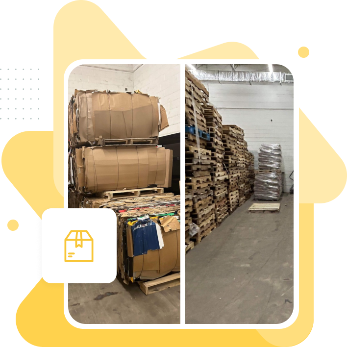 Stacked cardboard bales on pallets in warehouse, alongside a large pile of wooden pallets